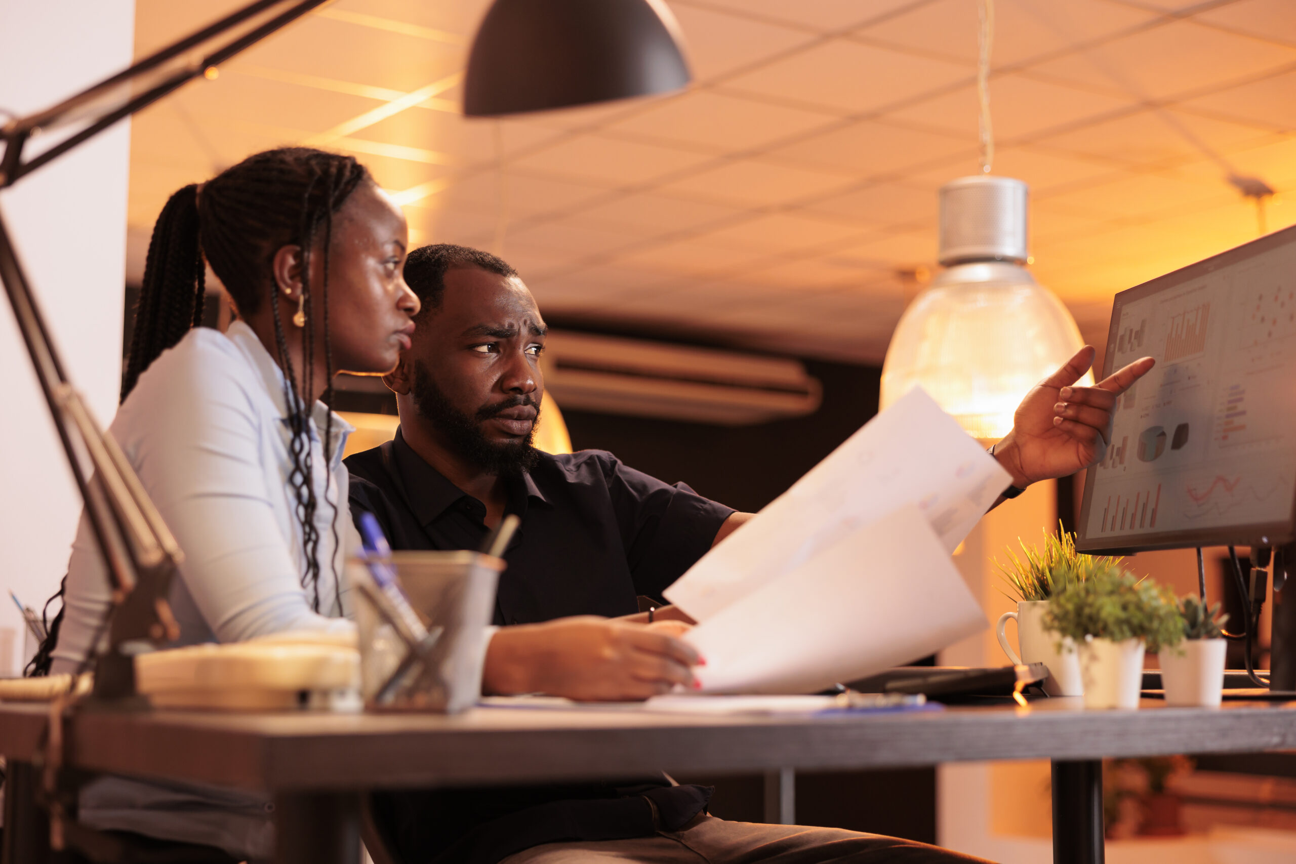 African american man and woman doing teamwork collaboration to do presentation report, looking at data on computer and papers. Working together with paperwork in office during sunset.